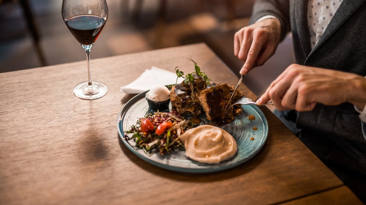 A man sitting at a wooden table cutting into a steak dinnr